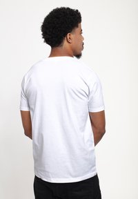 Man med kort lockigt hår och skägg, iklädd en enkel vit kortärmad t-shirt och svarta byxor, vänd bort mot en vit bakgrund.