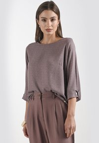 Bruine blouse met patroon, heeft een brede halslijn en driekwart mouwen. Gecombineerd met hooggehaalde bruine broeken met een geplisseerd ontwerp.