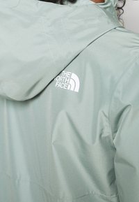 Mint green waterproof jacket with a hood, featuring a white logo on the upper back. Smooth texture and fitted design.