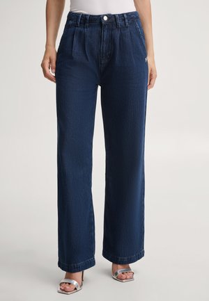 Donna che indossa jeans a vita alta, blu scuro, a gamba larga con pieghe, top bianco e sandali argentati con tacco, in piedi contro uno sfondo semplice.