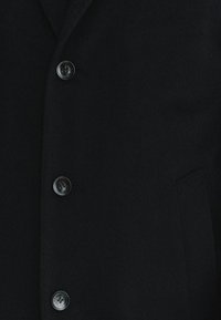 Czarny wełniany płaszcz z pięcioma zapięciami na guziki, klapami, oraz jedną boczną kieszenią o gładkiej w dotyku fakturze.