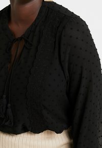 Sort bluse med prikket tekstur, bølget blondekant, bindebånd med frynser ved halsen og lange ærmer. Stoffet har en blød og let kvalitet.