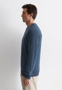 Blauer gerippter Pullover aus texturiertem Material, mit Rundhalsausschnitt und engen Bündchen, getragen über hellen Hosen.