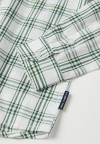 Valkoinen puuvillapaita, jossa on vihreä ruutukuvio, napitettu hihansuu ja tummansininen brändilabeli. Sileä pinta, jossa on tarkat viivat ja yksityiskohdat.