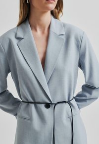 Svijetloplava sako jakna s prilagođenim krojem, oblikovanim reverima, jednim gumbom za zatvaranje i crnim remenom koji naglašava struk. Tkanina ima glatku teksturu.