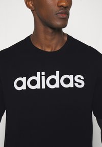Melna kokvilnas T-krekls ar lielu baltu "adidas" logo. Īsas piedurknes un apaļa kakla izgriezums. Gluda tekstūra ar minimālu apdari.