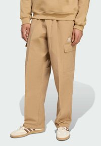 Pantalones cargo beige con una tela texturizada, que cuentan con un bolsillo lateral y el logotipo de Adidas en blanco. Combinados con zapatos blancos y marrones.