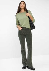 Maglione verde a maglia con una vestibilità rilassata, abbinato a pantaloni in velluto a coste verde scuro a palazzo. Il modello tiene una borsa nera in pelle e indossa scarpe nere.