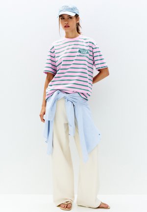 Stribet pink og grøn t-shirt med grafik, lys blå skjorte knyttet om taljen, cremefarvede brede bukser og afslappede sandaler.