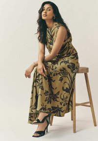 Rochie maxi din material verde măsliniu cu modele florale negre, purtată cu sandale cu toc înalt negre, așezată pe un scaun de lemn.