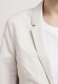 Vaalea beige puku takki, jossa on rakenteellinen kaulus ja lovetut läpät, tarjoamassa sileää pintaa. Päälle puettuna teksturoitu valkoinen toppi.