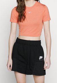 Osoba oblečená v korálovém zkráceném tričku Nike a černých šortkách Nike Air s bočními síťovanými panely na bílém pozadí.