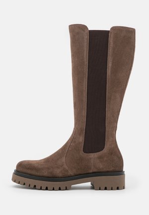Botte haute en daim marron avec panneau latéral élastique noir et semelle épaisse à crampons, vue de l'extérieur sur fond blanc.