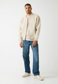 Giacca bomber beige con polsini a costine, indossata sopra una camicia beige chiara, abbinata a jeans azzurri a gamba dritta e sneakers bianche.