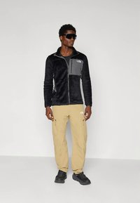 Hombre con chaqueta de forro polar negra, pantalones cargo beige, zapatillas negras y grandes gafas de sol negras, de pie contra una pared gris lisa.