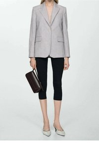 Mujer vistiendo un blazer gris claro, leggins negros cortos, tacones blancos de punta, sosteniendo un bolso marrón oscuro.