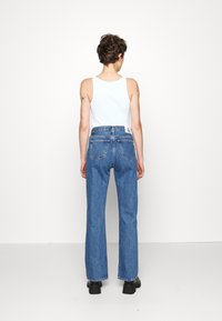 Blauwe denim jeans met een recht pijpdesign, hoge taille en zichtbare achterzakken. Draag met een witte tanktop, gezien van achteren.