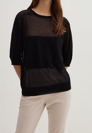 Kvinde iført en sort gennemsigtig strikket top med trekvartærmer og beige bukser, stående med hænderne bag ryggen.