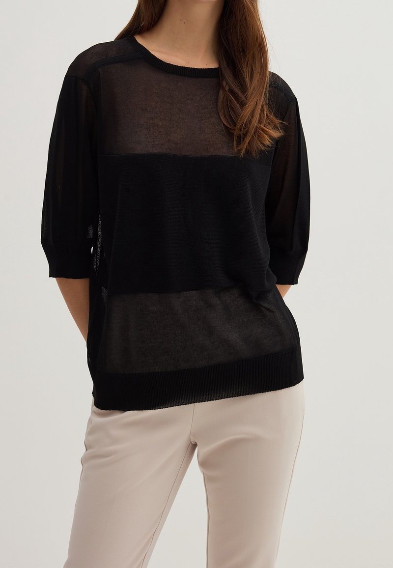 Femme portant un haut en maille noire transparente à manches trois-quarts et un pantalon beige, debout avec les mains dans le dos.
