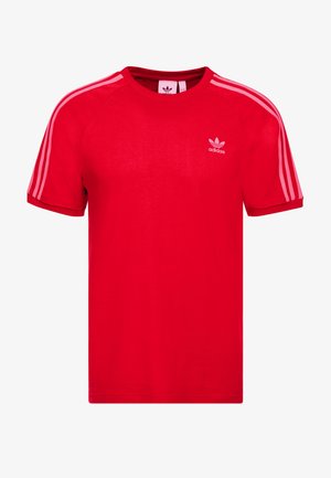 Czerwona koszulka Adidas z krótkim rękawem, z jasnoróżowymi trzema paskami na ramionach oraz małym logiem Adidas na lewej piersi.