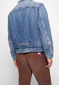 Denimjacka med en blekt blå tvätt, knäppning med knappar och en fårskinnskrage. Matchad med bruna kordfläskbyxor och en synlig varumärkesetikett.