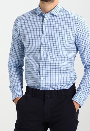 Homme portant une chemise bleu clair boutonnée avec un motif à carreaux bleu marine et un pantalon noir, mains dans les poches, sur un fond uni.