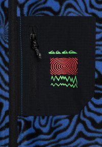 Vêtement en polaire avec un motif tourbillon bleu et noir. Présente une poche zippée noire avec des designs graphiques rouges et verts.