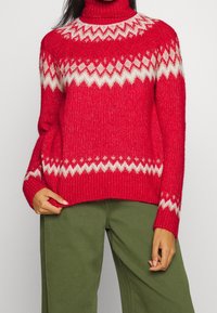 Red knitted sweater with a high turtleneck, featuring a white geometric pattern. Ribbed texture and long sleeves. Paired with green pants.