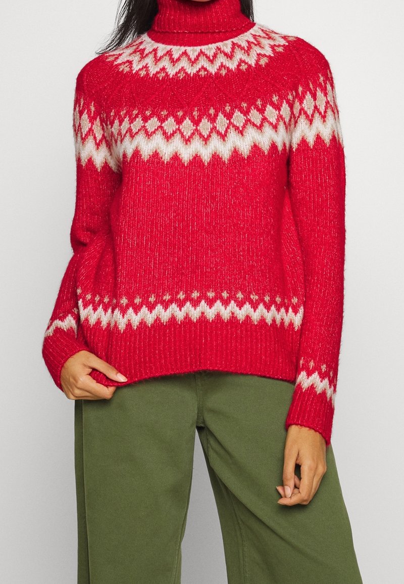 Red knitted sweater with a high turtleneck, featuring a white geometric pattern. Ribbed texture and long sleeves. Paired with green pants.