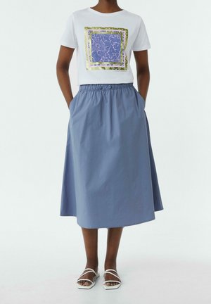 Frau trägt ein weißes T-Shirt mit blauem und goldenem quadratischem Design, einen blauen Midirock mit elastischem Bund und weiße flache Sandalen mit Riemchen.