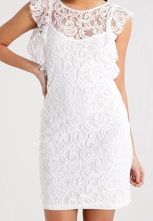 Robe blanche sans manches en dentelle avec des motifs floraux complexes et des manches courtes volantées, silhouette ajustée, portée par une personne avec une main sur la hanche.