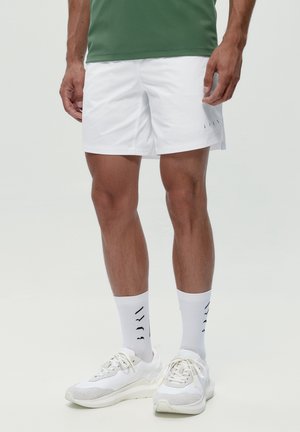 Hombre vistiendo pantalones cortos atléticos blancos, calcetines blancos con texto en negro, zapatillas blancas y una camiseta verde visible desde la cintura hacia abajo.