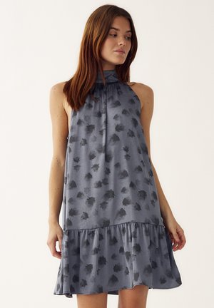 Žena s rovnými hnedými vlasmi, oblečená v sivých bezrukávových šatách s tmavým kvetinovým vzorom a riaseným lemom, otočená mierne doľava.