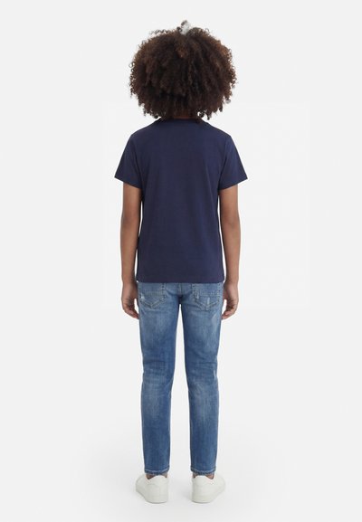 Enfant aux cheveux bouclés, de dos, portant un t-shirt bleu marine, un jean bleu délavé et des baskets blanches sur fond blanc.