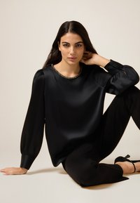 Top de satén negro de manga larga con escote redondeado, adornado con un detalle texturizado, combinado con pantalones negros ajustados y zapatos de tacón alto.