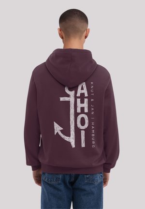 Person wearing a dark purple hoodie with a white anchor and "AHOI" text printed vertically on the back, paired with blue jeans.