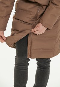Brun pufferjakke med lynlåsfortil og justerbar kant, parret med mørke jeans. Stoffet ser glat og polstret ud til isolering.