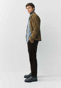 Young man standing sideways wearing olive suede jacket, light blue shirt, dark corduroy pants, and black shoes against plain background.
