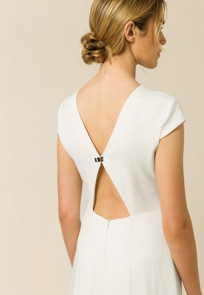 Robe blanche avec un dos en V profond et des manches courtes. Présente un petit fermoir décoratif à l'ouverture et une texture subtile. Fermeture éclair à la taille.