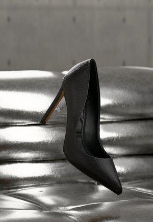 Chaussure à talon haut noire texturée, en équilibre sur son talon sur une surface métallique argentée brillante avec un fond uni flou.