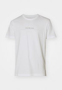 Camiseta de algodón blanca de manga corta. Cuenta con un cuello redondo y texto impreso en gris que dice "Calvin Klein Jeans" y "establecido en mil974."