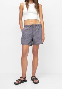 Nainen, joka on pukeutunut harmaisiin kiristysnauhoilla varustettuihin shortseihin, valkoiseen lyhyeen toppiin ja mustiin köysisaappaisiin, seisoo kädet taskuissa valkoista taustaa vasten.