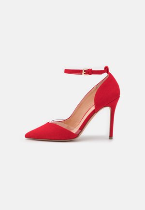 Escarpin rouge en daim avec un bout pointu et un cou-de-pied transparent, doté d'une bride réglable à la cheville avec une boucle en métal doré.