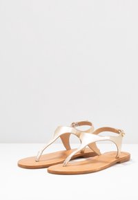 Kultaiset metallinväriset sandaalit, joissa on thong-muotoilu, litteä ruskea pohja ja nilkkaremmi. Sileä pinta, minimalistinen tyyli ja ei näkyviä kuvioita.