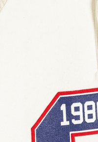 Camiseta de algodón blanca con un logo octogonal en azul marino y rojo con el número "1980" en blanco, sobre un fondo de tela texturizada.