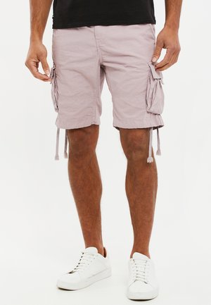 Helle rosa Cargo-Shorts mit Seitentaschen, Kordelzügen am Saum und einer lockeren Passform, gefertigt aus einem leichten Stoff. Getragen mit weißen Turnschuhen.
