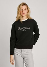 Sudadera negra con texto blanco "Pepe Jeans London", escote redondo, mangas largas, puños y dobladillo acanalados. Llevada con jeans gris claro.