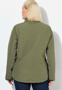 Olijfgroene jas met een gequilt herringbone patroon, hoge kraag en aansluitende mouwen. Gladde textuur en zachte stof.
