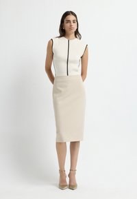 Ärmelloses, geripptes Oberteil in Creme mit schwarzem Saum, kombiniert mit einem beigen knielangen Bleistiftrock und beigen, spitzen High Heels. Glatter Stoff. 
