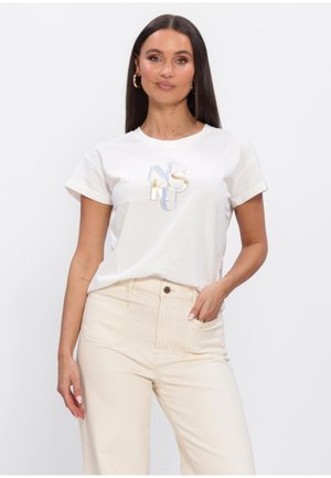 Vrouw met lang donker haar, die gouden oorringen draagt, een wit t-shirt met versierd logo en een high-waisted crème broek met hand in de zak.